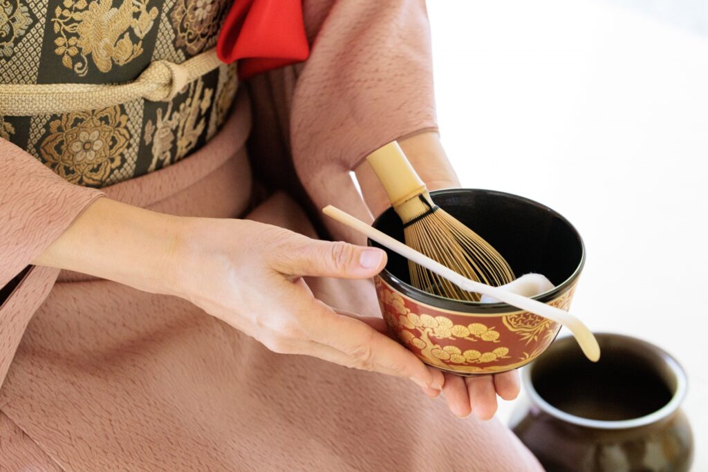 着物姿の人物が茶碗と茶筅・茶杓を丁寧に持っている様子。裏千家の茶道具を象徴する品格ある構成。